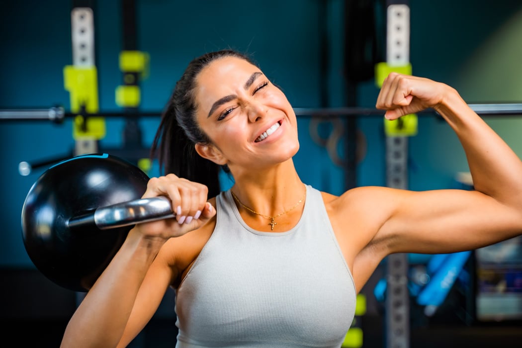 Coach is holding a kettlebell on her right hand and flexing her left one.